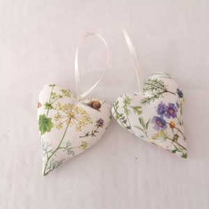 May include: Two heart-shaped fabric ornaments featuring a white background with a botanical print of yellow and purple wildflowers, green foliage, and a small brown snail. Each heart includes a thin white ribbon loop for hanging purposes.
