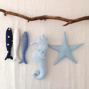 May include: A collection of nautical-themed fabric ornaments hanging from a brown branch. The ornaments include fish, a seahorse, and a starfish, all in shades of blue and white. The fish have polka dot and striped patterns. The seahorse and starfish have a gingham and polka dot pattern.