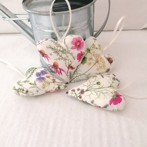 May include: Three heart-shaped fabric ornaments featuring a botanical print of pink cosmos, yellow wildflowers, and green foliage. Each heart includes a thin white ribbon loop for hanging. The ornaments are arranged in front of a galvanized metal watering can.