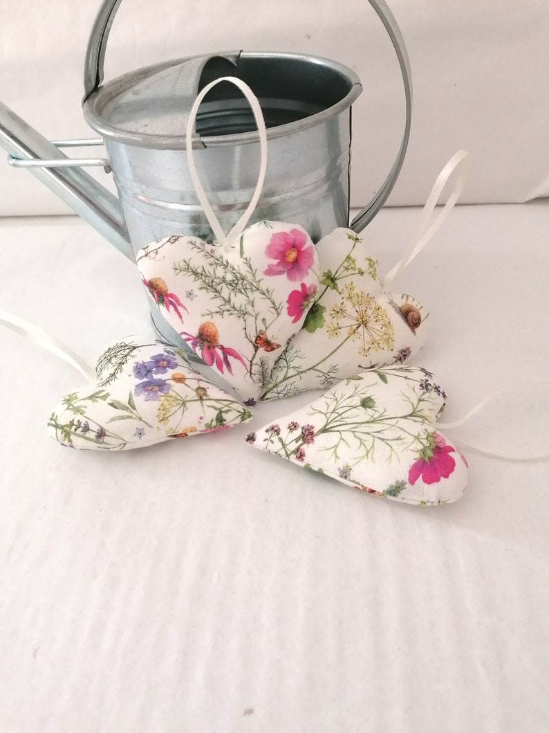 May include: Three heart-shaped fabric ornaments featuring a botanical print of pink cosmos, yellow wildflowers, and green foliage. Each heart includes a thin white ribbon loop for hanging. The ornaments are arranged in front of a galvanized metal watering can.