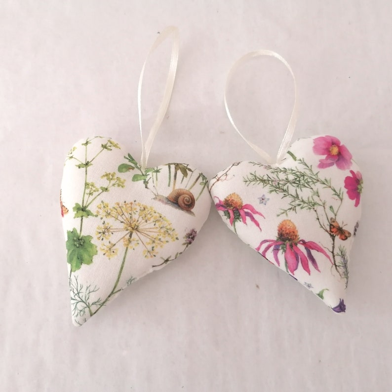 May include: Two heart-shaped fabric ornaments featuring a white background with botanical illustrations of yellow and pink flowers, green foliage, a small brown snail, and a butterfly. Each heart includes a thin white ribbon loop for hanging.
