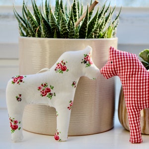 May include: Two fabric toy horses, one white with pink flowers and one red and white gingham. The horses are sitting on a white surface in front of a potted succulent plant.