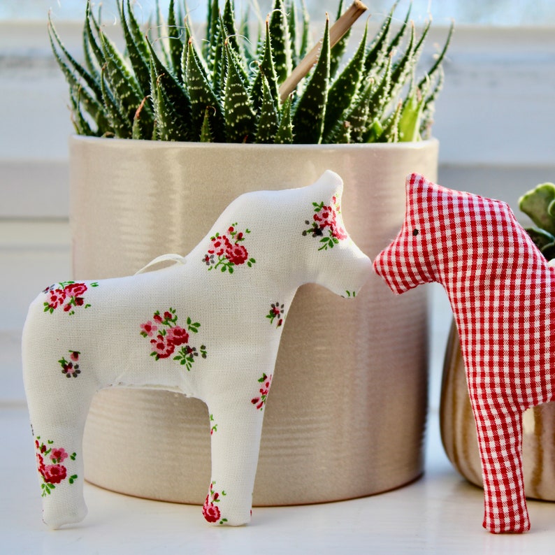 May include: Two fabric toy horses, one white with pink flowers and one red and white gingham. The horses are sitting on a white surface in front of a potted succulent plant.