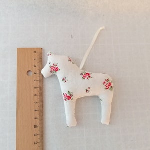 May include: A white fabric Dala horse ornament with a floral pattern in shades of pink and green. The horse is approximately 5 inches tall and has a white ribbon for hanging.