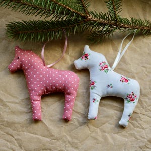 May include: Two handmade fabric horse ornaments. One is pink with white polka dots and the other is white with a floral pattern. Both have a white ribbon for hanging.