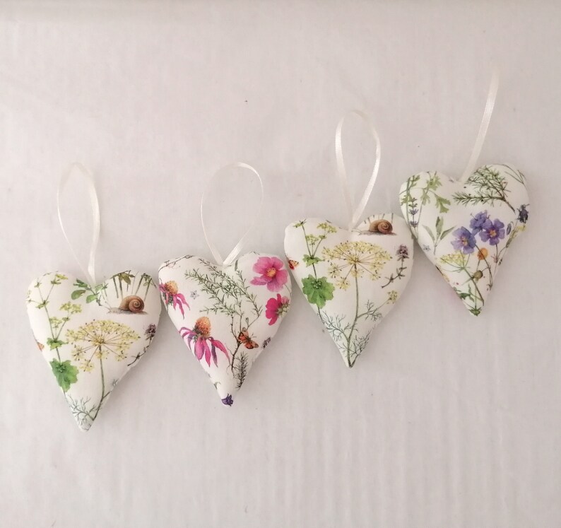 May include: Four heart-shaped fabric ornaments featuring a white background with botanical illustrations of wildflowers, green foliage, small insects, and snails. Each heart includes a thin white ribbon loop attached to the top for hanging purposes.