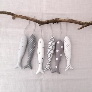 May include: Seven fabric fish ornaments in various shades of gray and white hang from a brown branch. The fish have different patterns, including polka dots, stars, and a wave design. Each fish is suspended by a thin string.