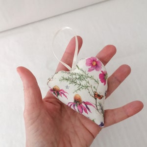 May include: A heart-shaped fabric ornament featuring a white background with a botanical print of pink coneflowers, green foliage, and a small orange butterfly. The ornament includes a thin white ribbon loop attached to the top for hanging.