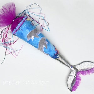Könnte beinhalten: Ein blauer und weißer Stoffkegel mit einer daran befestigten Meerjungfrauenflosse. Der Kegel ist mit Delfinen und Muscheln verziert. Die Flosse ist silberfarben mit rosa Tüllbesatz. Der Kegel ist mit einer rosa Tüllschleife verziert.