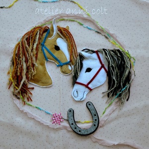 Könnte beinhalten: Applizierte Pferdsköpfe mit Garnmähnen und blauen und roten Zäumen. Ein Hufeisen und eine rosa Blume sind ebenfalls im Design enthalten. Der Text "atelier anni colt" steht in der oberen linken Ecke. Der Hintergrund ist ein hellrosa Stoff.