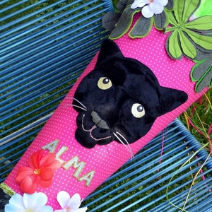 Könnte beinhalten: Eine rosa Schultüte mit weißen Punkten, einem schwarzen Panthergesicht und dem Namen "ALMA" in silbernen Glitzerbuchstaben. Verziert mit Stoffblumen und Blättern, liegt sie auf einem blauen Metallstuhl.