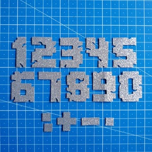 Könnte beinhalten: Silberne Glitzerzahlen und -symbole auf einer blauen Rasteroberfläche. Die Zahlen 1 bis 0 werden angezeigt, zusammen mit einem Pluszeichen, einem Minuszeichen, einem Punkt und einem Doppelpunkt. Die Zahlen und Symbole sind pixelig.