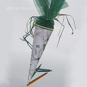 Könnte beinhalten: Eine graue, kegelförmige Schultüte mit einem naturwissenschaftlichen und mathematischen Muster, einschließlich Fliegen, dem Pi-Symbol und Leiterplattendesigns. Die Tüte ist mit grünem Tüll und Bändern verziert. Der Text "atelier anni colt" ist sichtbar.
