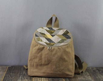 Könnte beinhalten: Ein brauner Canvas-Rucksack mit einer geometrischen Musterklappe. Die Klappe besteht aus einem Stoff mit schwarzen, weißen und goldenen geometrischen Formen.
