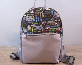 Könnte beinhalten: Ein grau-schwarzer Rucksack mit einem Gamer-Thema. Der Rucksack hat ein großes Frontfach und verstellbare Träger. Der Stoff zeigt ein sich wiederholendes Muster aus Videospielcontrollern, Wörtern wie "Start", "Play", "Level" und "Score" sowie farbenfrohe Pixelgrafiken.