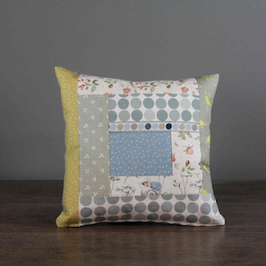 Könnte beinhalten: Ein Patchwork-Kissen mit quadratischem Design, das verschiedene Muster in Blautönen, Grün- und Gelbtönen zeigt. Das Kissen hat einen weißen Hintergrund mit kleinen Blumenmustern und Punkten.