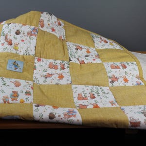 Könnte beinhalten: Eine gelbe und weiße Patchwork-Decke mit einem verspielten Tierdruck. Die Decke besteht aus Quadraten aus verschiedenen Stoffen, die jeweils ein anderes Tierdesign aufweisen. Die Decke ist in der Hälfte gefaltet und liegt auf einer Holzoberfläche.