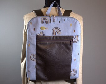 Könnte beinhalten: Ein hellblauer Rucksack mit einer braunen Kunstleder-Vordertasche. Der Rucksack hat ein Muster aus goldenen Regenbögen und braunen Regentropfen.