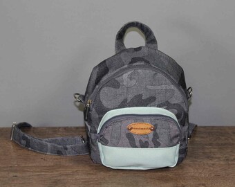 Könnte beinhalten: Ein kleiner grauer und grüner Tarnrucksack mit einem hellgrünen Boden und einem braunen Lederpatch mit der Aufschrift "handgefertigt".