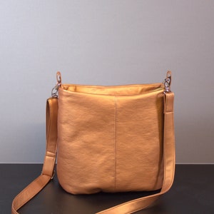 Könnte beinhalten: Eine metallisch goldfarbene Schultertasche mit einem langen, verstellbaren Trageriemen. Die Tasche hat ein schlichtes, elegantes Design und ist aus einem glatten, strukturierten Material gefertigt.