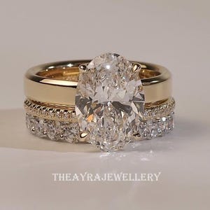 Könnte beinhalten: Ein dreiteiliges goldenes Ringset. Der Verlobungsring hat einen großen, oval geschliffenen Diamanten. Der Ehering ist ein schlichtes Goldband, und das zweite Band ist mit kleineren Diamanten besetzt. Der Text "THEAYRAJJEWELLERY" befindet sich unten.
