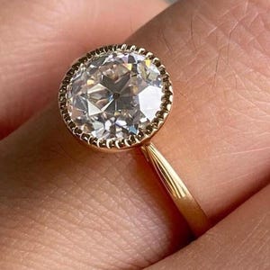 May include: A gold ring featuring a large, round, clear diamond with a detailed, beaded bezel setting. The ring's band is a simple, polished gold. The diamond sparkles with many facets.