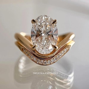 May include: A gold engagement ring featuring a large oval-cut diamond. The ring is accompanied by a curved band adorned with small diamonds. The ring is displayed on a reflective surface. The text "THEAYRAJJEWELLERY" is visible.