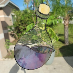 Può includere: Bottiglia di pozione in vetro colorato con un liquido iridescente viola. La bottiglia ha un tappo giallo ed è bordata di nero. Una catena d'argento è attaccata per appenderla. Lo sfondo mostra fogliame verde e cielo blu.