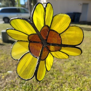 May include: A stained glass sunflower suncatcher with yellow petals and a brown center. The glass is outlined in black and hangs from a silver chain. The suncatcher is displayed outdoors in natural light.