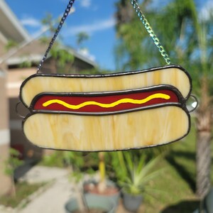 May include: A stained glass hot dog, featuring a yellow bun, red sausage, and yellow mustard. The glass has a yellow and white marbled effect, with black outlines. A decorative chain is attached for hanging.