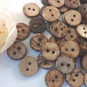 Könnte beinhalten: Eine Sammlung brauner Kokosnuss-Knöpfe mit jeweils zwei Löchern. Die Knöpfe sind auf einer weißen Oberfläche verstreut.