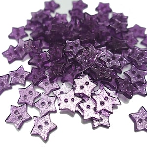 May include: A large pile of purple star-shaped buttons with a glittery finish. The buttons are scattered on a white background.