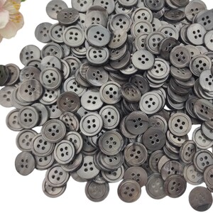 Könnte beinhalten: Ein großer Haufen grauer, vierlöchriger Knöpfe. Die Knöpfe sind aus einem glatten, glänzenden Material gefertigt und haben einen leicht erhöhten Rand.
