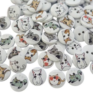 May include: A collection of white wooden buttons featuring a variety of cat designs. The buttons are round and have two holes. The cats are shown in different poses and colours.