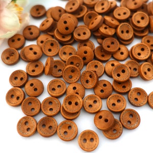 Könnte beinhalten: Ein großer Haufen brauner Holzknöpfe mit jeweils zwei Löchern. Die Knöpfe haben einen genähten Rand und sind auf einer weißen Oberfläche verstreut.