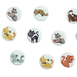 Pode incluir: Conjunto de 12 botões de madeira com vários desenhos de gatos. Os botões são brancos com ilustrações coloridas de gatos. Os gatos são representados em várias poses, incluindo dormir, brincar e sentar.