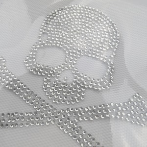 Könnte beinhalten: Ein weißer Stoffhintergrund mit einem Totenkopf- und Knochenkreuz-Design aus silbernen Strasssteinen.