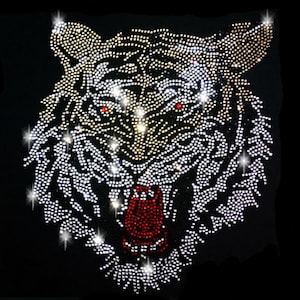 Può includere: Un design di testa di tigre bianca realizzato con strass su uno sfondo nero. Gli occhi del tigre sono rossi e la bocca è aperta, mostrando denti rossi.
