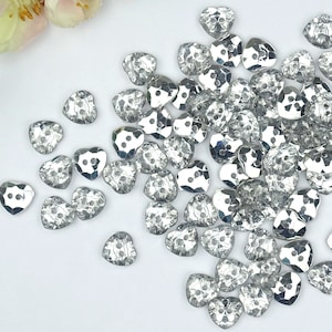 May include: A large quantity of silver heart-shaped buttons. The buttons are faceted and have a shiny, reflective finish.