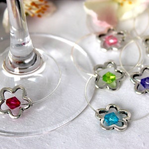 Op de afbeelding: Wijnglasbedels met bloemvormige zilverkleurige metalen frames en kleurrijke kristallen centra. De bedels zijn bevestigd aan dunne, transparante draden. De kristallen zijn rood, roze, groen, geel, blauw en paars.