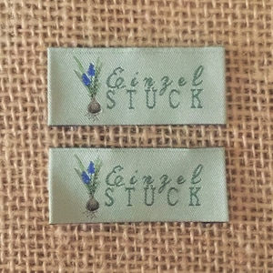 Könnte beinhalten: Zwei grüne Stoff-Etiketten mit weißem Text "Einzel Stück" und einer kleinen blauen Blumengrafik.