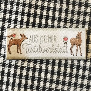 Könnte beinhalten: Ein weißes Stofflabel mit dem Text "AUS MEINER Textilwerkstatt" in grauer Schrift. Das Label zeigt zwei braune Rehe, eines mit einem roten Rotkehlchen auf dem Rücken, und einen roten Kreis mit einem weißen Punkt in der Mitte.