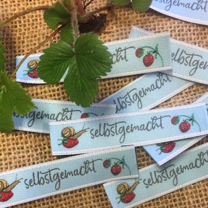 Könnte beinhalten: Eine Sammlung von blauen und weißen Stoff-Etiketten mit dem Text "selbstgemacht" und einer kleinen Illustration einer Schnecke und einer Erdbeere.