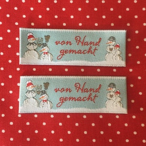 Könnte beinhalten: Zwei weiße Stoff-Etiketten mit blauen Rändern und einem Schneemann-Motiv. Die Etiketten lauten "von Hand gemacht", was "handgemacht" bedeutet.