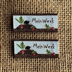 Könnte beinhalten: Zwei braune und weiße gewebte Etiketten mit einem Cartoon-Bild eines Maulwurfs, der im Boden gräbt, mit einer grünen Pflanze und einem roten Rettich. Der Text auf dem Etikett lautet "Mein Werk".