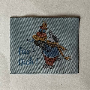 Könnte beinhalten: Ein blauer Stoff-Patch mit einem Comic-Dachs, der einen rot-weißen Hut trägt und einen Stapel Geschenke hält. Der Text "Für dich!" ist auf dem Patch aufgedruckt.