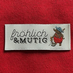 Könnte beinhalten: Ein rechteckiges weißes Etikett mit den Worten "fröhlich & MUTIG" in schwarzer Schrift und einem Cartoon-Bären in roter Latzhose und blauem Hut. Das Etikett befindet sich auf rotem Stoff.