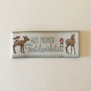 May include: White fabric label with the text "AUS MEINER TEXTILWERKSTATT" in silver lettering. The label features two brown deer, one with a red robin perched on its back.