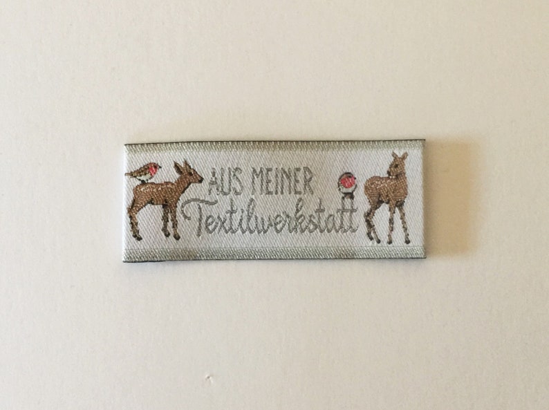 May include: White fabric label with the text "AUS MEINER TEXTILWERKSTATT" in silver lettering. The label features two brown deer, one with a red robin perched on its back.