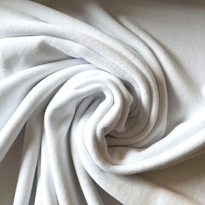 May include: Close-up of a white fabric with a soft, velvety texture. The fabric is draped in gentle folds, creating shadows and highlights that emphasize its plushness. The material appears to be a smooth, high-quality textile, suitable for various sewing projects.